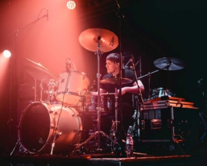 A drummer performing on stage with vibrant lighting effects.